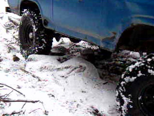 1985 Ford Bronco - The side of a blue 1985 Ford Bronco is shown with its large tires on a snowy, uneven terrain. The vehicle appears to be off-roading.