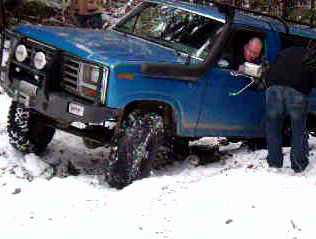1985 Ford Bronco - A blue 1985 Ford Bronco is stuck in the snow with its front tires turned. A person is visible in the driver's seat and another person is st…
