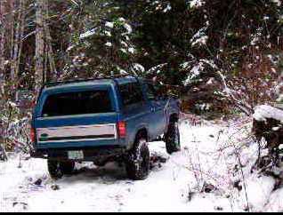 1985 Ford Bronco - The rear-left quarter view of a blue Ford Bronco is shown driving on a snowy, wooded trail. The vehicle has off-road tires and appears to b…