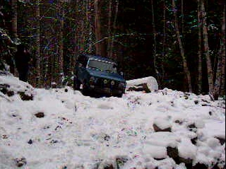 1985 Ford Bronco - A blue 1985 Ford Bronco is navigating a snow-covered, wooded trail. The vehicle is angled towards the viewer, showing its front and driver'…