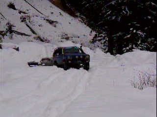 1985 Ford Bronco - A blue 1985 Ford Bronco is driving through deep snow, pulling a trailer behind it on a snowy mountain road.