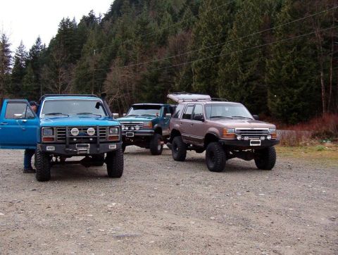 1985 Ford Bronco - Three modified off-road SUVs are parked on a gravel lot with a wooded hillside in the background. The closest vehicle is a blue Ford Bronco…
