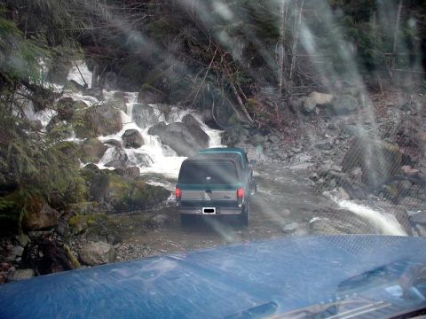 1985 Ford Bronco - A dark green Ford Bronco is driving through a shallow stream with a waterfall in the background. The photo is taken from the perspective of…