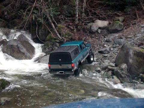 1985 Ford Bronco - A teal pickup truck with a black camper shell is driving through a rocky riverbed. The truck is angled towards the right, with its rear fac…