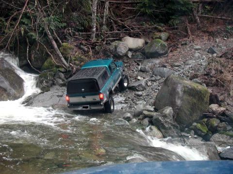 1985 Ford Bronco - A teal Ford Bronco is navigating a rocky stream bed with a waterfall to its left. The truck is equipped with a camper shell and appears to…