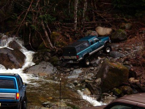 1985 Ford Bronco - A teal Ford Ranger pickup truck with a camper shell navigates a rocky, wooded terrain next to a rushing stream. Another blue SUV is partial…