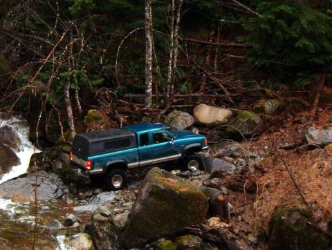 1985 Ford Bronco - A teal pickup truck with a black camper shell navigates a rocky stream bed in a wooded area.