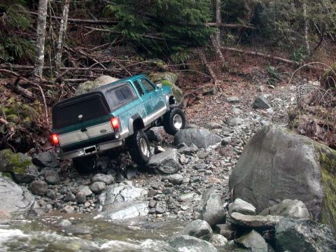 1985 Ford Bronco - A teal Ford Bronco with a black camper shell navigates a rocky stream bed in a wooded area. The vehicle is angled with its rear facing the…