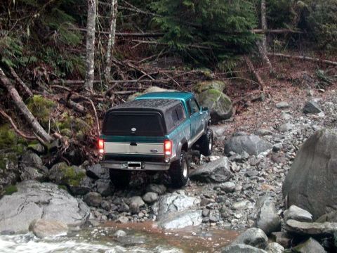 1985 Ford Bronco - A teal Ford pickup truck with a black camper shell navigates a rocky, wooded terrain next to a shallow stream. The truck is viewed from the…