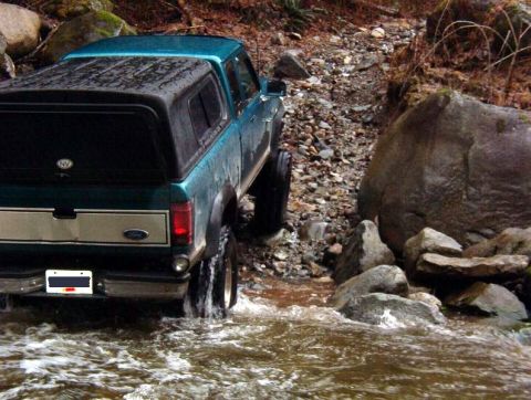 1985 Ford Bronco - The rear driver's side of a teal Ford pickup truck with a black camper shell is driving through shallow, rocky water.
