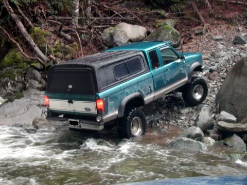 1985 Ford Bronco - A teal Ford Ranger pickup truck with a black camper shell navigates through a rocky stream bed. The truck is angled to the right, with its…