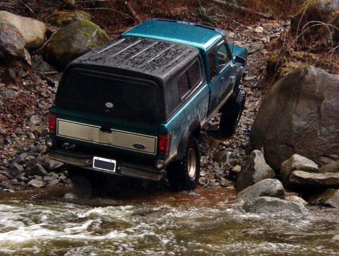 1985 Ford Bronco - A teal Ford pickup truck with a black camper shell drives through a rocky stream. The truck is angled towards the right side of the frame,…