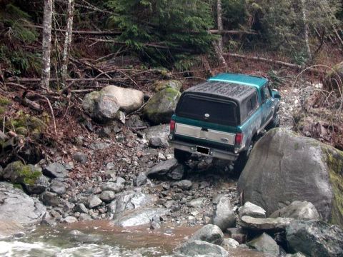 1985 Ford Bronco - A teal Ford pickup truck with a camper shell navigates a rocky, wooded stream bed.