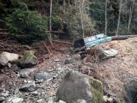 1985 Ford Bronco - A teal pickup truck with a camper shell is parked precariously on a steep, wooded embankment, with its rear wheels off the ground.