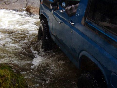 1985 Ford Bronco - A blue 1980s Ford Bronco is driving through a shallow river with rocky banks and mossy rocks. The tires are submerged in the fast-moving wa…