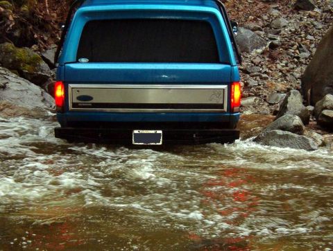 1985 Ford Bronco - The rear of a blue SUV is shown driving through a shallow, rocky stream. The vehicle's taillights are illuminated, and its license plate is…
