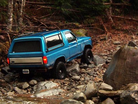 1985 Ford Bronco - A blue Ford Bronco navigates a rocky, wooded terrain next to a shallow stream. The rear of the SUV is visible, showing its taillights and a…