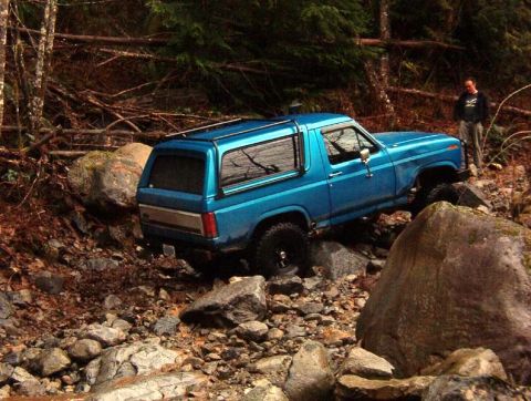 1985 Ford Bronco - A blue Ford Bronco is navigating a rocky, wooded terrain with a person standing in the background.