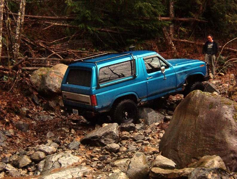 A blue Ford Bronco off-roading over rocks in a wooded area.