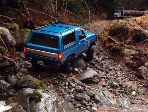 1985 Ford Bronco - A blue 1980s Ford Bronco is navigating a rocky, wet trail in a forest. Another vehicle is visible further up the trail.
