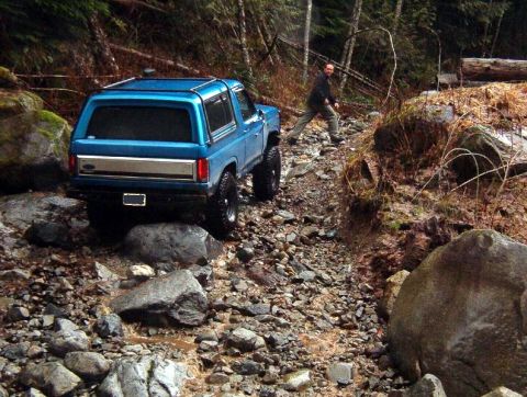1985 Ford Bronco - A blue Ford Bronco is navigating a rocky, wet trail in a wooded area. A person is visible walking behind the vehicle.
