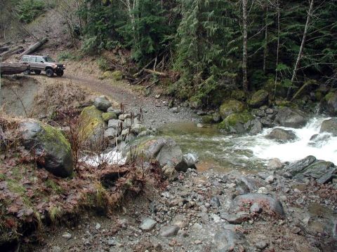 1985 Ford Bronco - A tan SUV with a camper shell is parked on a dirt road next to a rocky creek in a wooded area. The vehicle is positioned on an embankment o…