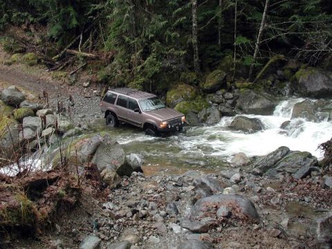 1985 Ford Bronco - A tan SUV is fording a rocky stream in a wooded area. The vehicle is angled towards the right, with its front tires submerged in the water.