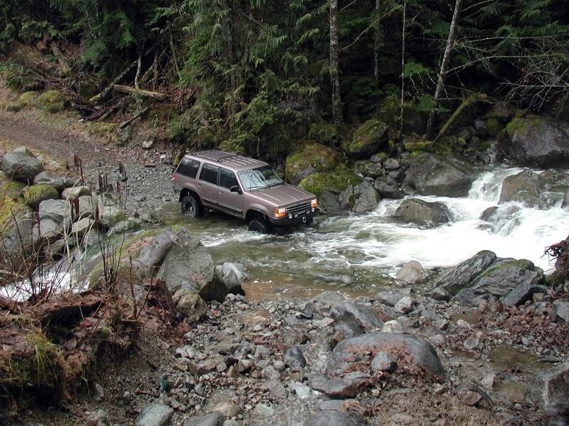Tan SUV fording a rocky stream in a wooded area.