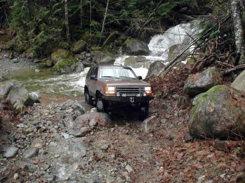 1985 Ford Bronco - A brown 1980s SUV navigates a rocky stream bed with a waterfall in the background. The vehicle has a modified front bumper with tow hooks a…