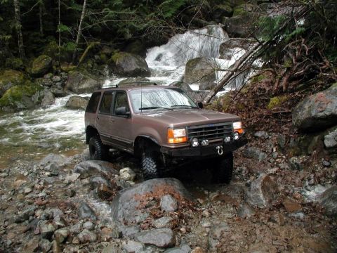 1985 Ford Bronco - A brown Ford Explorer is navigating a rocky stream bed with a waterfall in the background. The SUV has off-road tires and auxiliary lights…