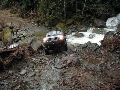1985 Ford Bronco - A brown 1985 Ford Bronco navigates a rocky, shallow stream crossing with a waterfall in the background.