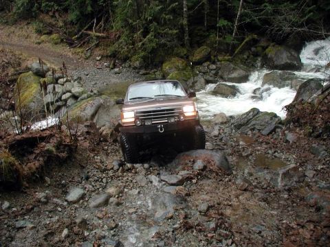 1985 Ford Bronco - A brown Ford Bronco navigates a rocky, wet trail next to a rushing stream. The vehicle is angled towards the camera, showing its front bump…