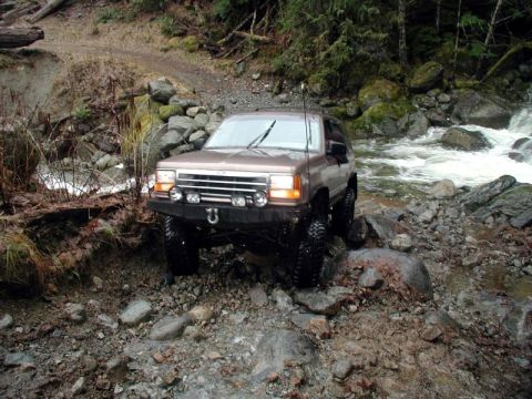 1985 Ford Bronco - A brown 1980s Ford SUV is navigating a rocky, off-road stream crossing with a fast-flowing river to its right.