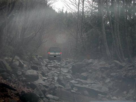 1985 Ford Bronco - A dark-colored SUV drives down a rocky, unpaved forest road. The vehicle is viewed from the rear, with its brake lights illuminated.