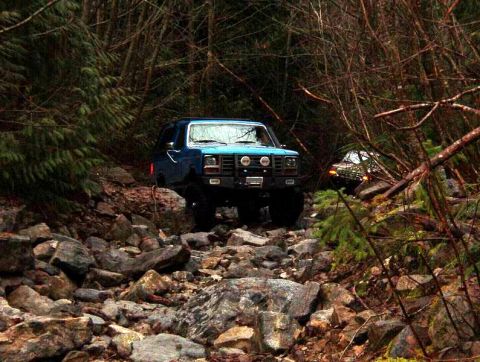 1985 Ford Bronco - A blue 1980s Ford Bronco is navigating a rocky, wooded trail. Another vehicle is visible further down the path.