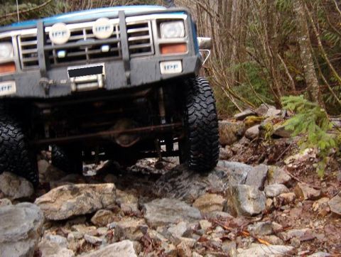 1985 Ford Bronco - The front end of a blue off-road SUV is angled over a rocky terrain, with large tires visible. The vehicle has a brush guard with auxiliary…