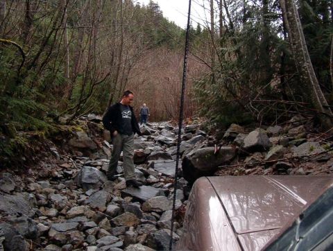 1985 Ford Bronco - The front right corner of a brown SUV is visible in the foreground, driving on a rocky, wooded trail. Two men are walking ahead on the trai…