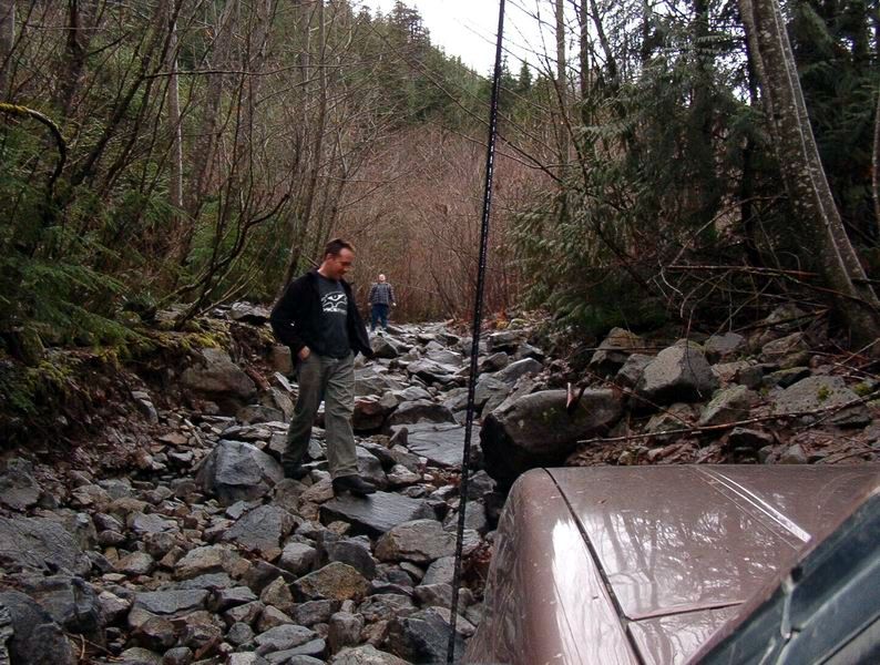 Brown SUV on a rocky trail in a forest with two men walking ahead.