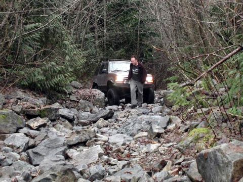 1985 Ford Bronco - A man walks next to a silver Ford Bronco as it navigates a rocky, wooded trail. The vehicle is positioned on a steep incline with large bou…