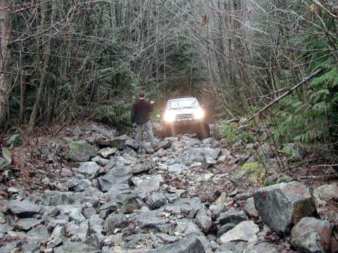 1985 Ford Bronco - A person stands on a rocky trail, pointing towards a white SUV driving through the dense forest. The SUV's headlights are on, illuminating…