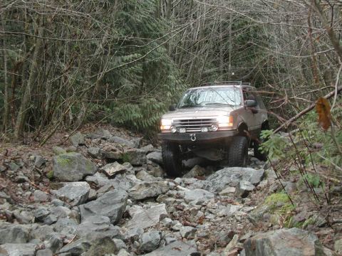 1985 Ford Bronco - A tan SUV navigates a rocky, wooded trail. The vehicle has a modified front bumper with a tow hook and aftermarket headlights.