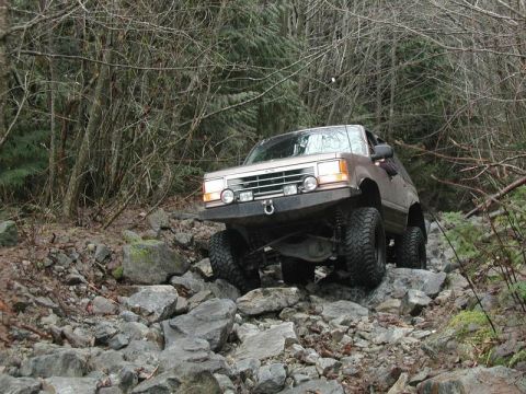 1985 Ford Bronco - A lifted, tan 1980s Ford Bronco navigates a rocky, wooded trail. The off-road vehicle has large tires and an aftermarket bumper with auxili…