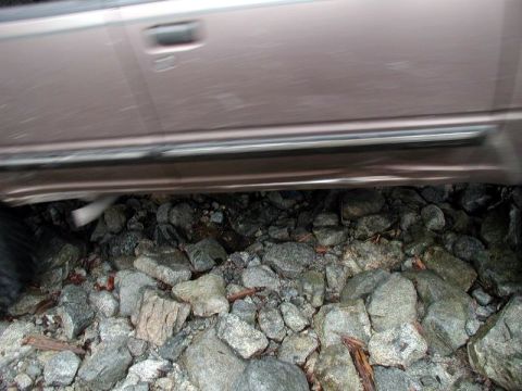 1985 Ford Bronco - The lower body of a tan SUV is visible, showing the door, running board, and exhaust pipe. The vehicle is parked over a rocky, uneven surfa…
