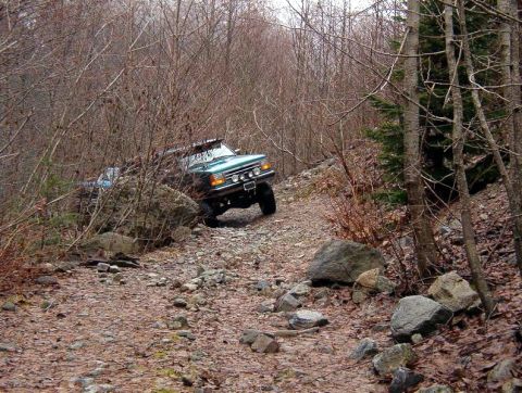 1985 Ford Bronco - A teal Ford Bronco is navigating a rocky, wooded trail. The vehicle is angled uphill, with its front end visible as it maneuvers around lar…