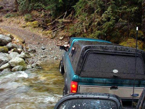 1985 Ford Bronco - A teal Ford pickup truck with a camper shell drives through a shallow stream on a rocky, wooded trail. The rear of the truck is visible, sh…