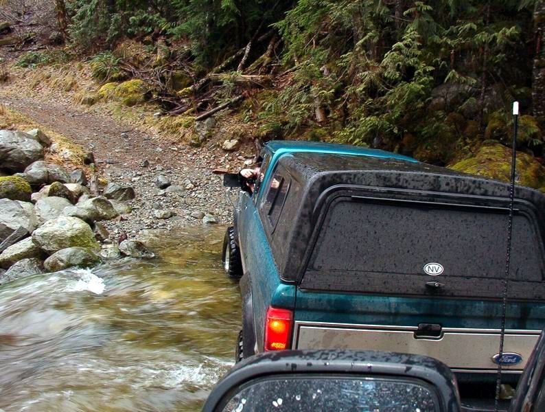 Teal Ford pickup truck with camper shell driving through a stream on a wooded trail.