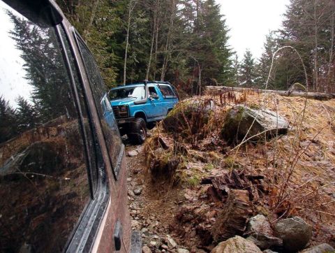1985 Ford Bronco - A blue 1980s Ford Bronco is navigating a rocky, wooded trail. The vehicle is viewed from the perspective of another vehicle, with its side…
