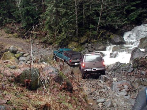 1985 Ford Bronco - Two off-road vehicles navigate a rocky stream bed with a waterfall in the background. A teal pickup truck and a brown SUV are shown from th…