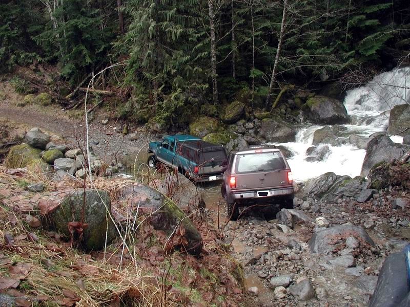 Teal pickup truck and brown SUV driving through a rocky stream bed.