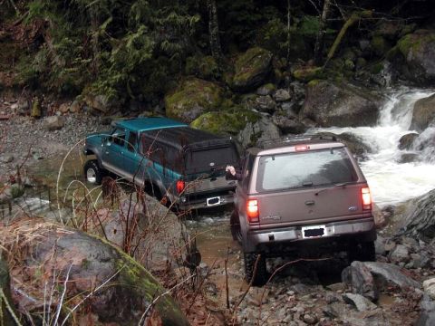 1985 Ford Bronco - Two off-road vehicles navigate a rocky stream bed with a waterfall in the background. A teal extended cab pickup truck with a camper shell…